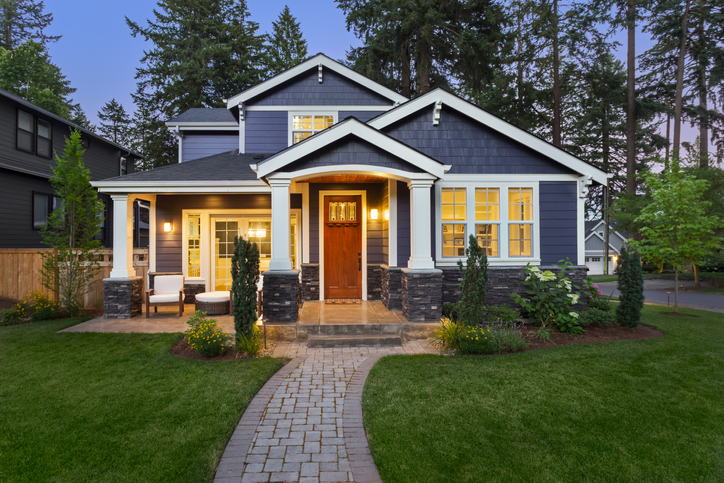 facade of home with manicured lawn, landscaping, and backdrop of trees and dark blue sky. Glowing interior lights create a welcoming mood.